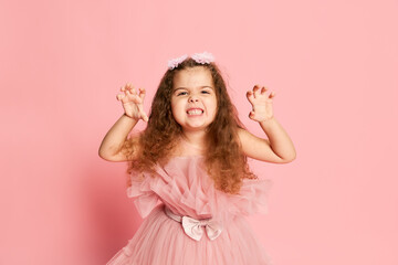 Child angry. Expressive little girl, kid with long curly hair wearing festive princess dress isolated over pink background. Concept of facial expression, childhood, child emotions