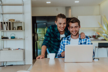 A young married couple is talking to parents, family and friends on a video call via a laptop while sitting in the living room of their modern house