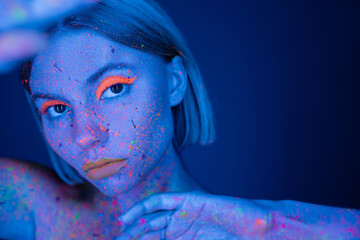portrait of woman with fluorescent makeup and colorful neon body paint on blurred foreground isolated on dark blue.