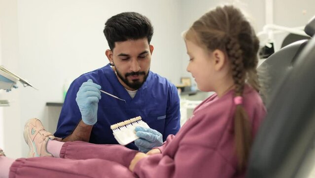 Young professional bearded male dentist choosing color of filling from palette for little female child in moderm dental clinic. Visual method of subjective perception color of filling by a doctor.