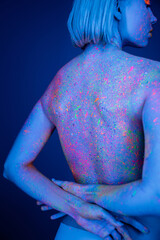 woman with body in bright neon splashes standing with hands behind back isolated on dark blue.