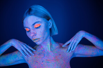 pretty woman in bright makeup and glowing neon body paint posing isolated on dark blue.