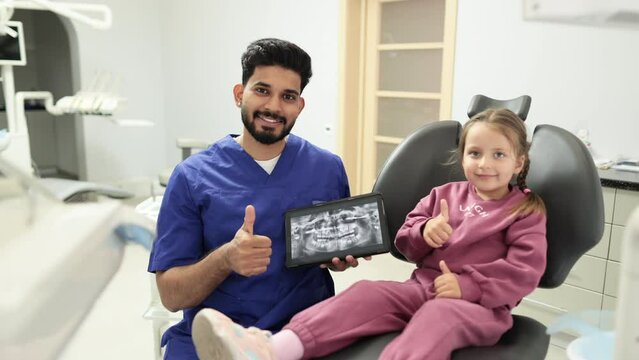 Smiling caucasian preschool girl, sitting on dentist chair and looking at digital tablet with x-ray scan image of teeth together with her professional male bearded dentist at clinic showing thumbs up.
