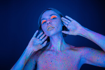 naked woman in neon makeup and body in glowing paint posing with hands near face isolated on dark blue.