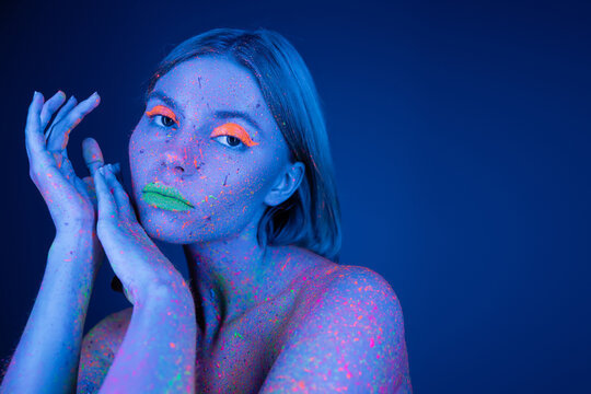 woman in vibrant makeup and colorful neon splashes on body looking at camera isolated on dark blue.
