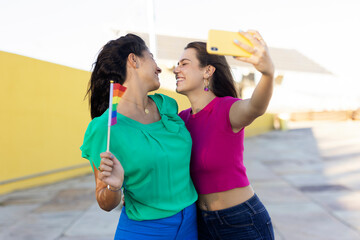 Beautiful lesbian young couple embraces and holds a rainbow flag. Girls taking selfie photo..