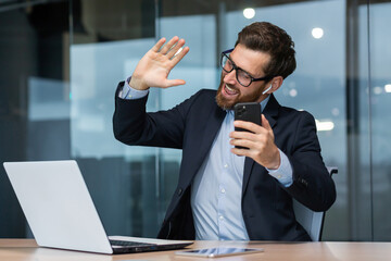 Young handsome man sitting in a suit in the office at the table and listening to music from the phone in headphones. Dances along, relaxed, waves his hands.