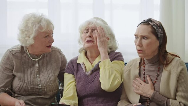 Three elderly women watching TV at home, feeling shocked with bad news, media