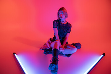 full length of woman in trendy boots sitting near purple neon lamps and looking away on coral background.