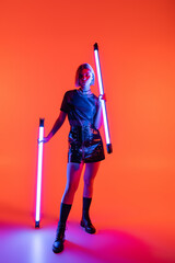 full length of woman in mini skirt holding bright neon lamps on purple and coral background.