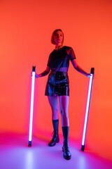 full length of slender and trendy woman holding neon lamps and looking away on coral and purple background.