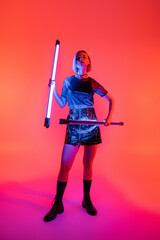 full length of trendy woman in leather boots and skirt holding neon lamps on purple and coral background.