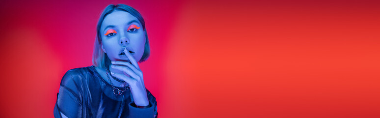 stylish woman with neon makeup touching lips in blue light on coral red background, banner.