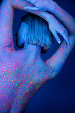 back view of nude woman colored with neon paint posing with hands behind head isolated on dark blue.
