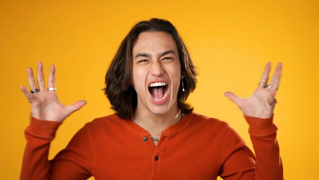 Portrait of angry, frustrated upset Hispanic Latino gender fluid man yelling with clenched fists isolated on yellow background.