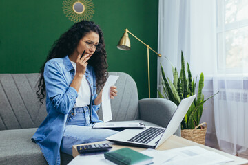 Serious and thoughtful businesswoman working at home with documents, hispanic woman in casual clothes using laptop at work sitting on sofa in living room with bills and contracts.