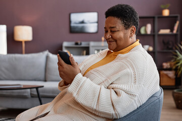 Side view portrait of senior black woman using smartphone while relaxing in chair in cozy home setting and smiling cheerfully