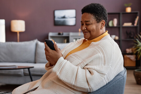 Side View Portrait Of Senior Black Woman Using Smartphone While Relaxing In Chair In Cozy Home Setting And Smiling Cheerfully