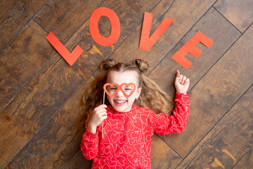 a little girl child in red pajamas with the inscription Love lies on a dark brown wooden background on the floor and laughs holding glasses in the form of hearts, the concept of Valentine's day