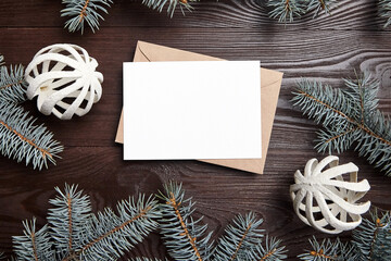 Christmas greeting card mockup with envelope, green fir tree branches and decorations on brown wooden background, top view, flat lay. White New Year holiday card with winter decor