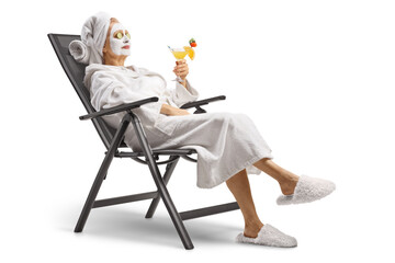 Woman in a bathrobe with a face mask and cucumber on eyes relaxing with a cocktail on a chair