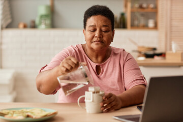 Fototapeta na wymiar Warm toned portrait of black senior woman making tea during breakfast in cozy kitchen