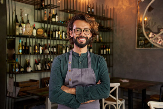 Looking at camera portrait of a muslim owner of a small local business. Cheerful male bartender smiling with positive attitude. Cheerful bar worker smiles while standing indoors.