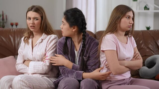 Young Asian woman talking to argued Caucasian friends forcing ladies making peace shaking little fingers. Satisfied happy lady smiling as women gesturing pinky swear sitting on couch in pajamas