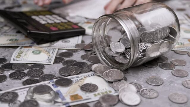Woman Counting Expenses on a Calculator on Paper in Front of a Jar with Coins. Ukrainian cents and 100-dollar bills are scattered on the table. Budget, payment, spending, savings. Financial crisis.