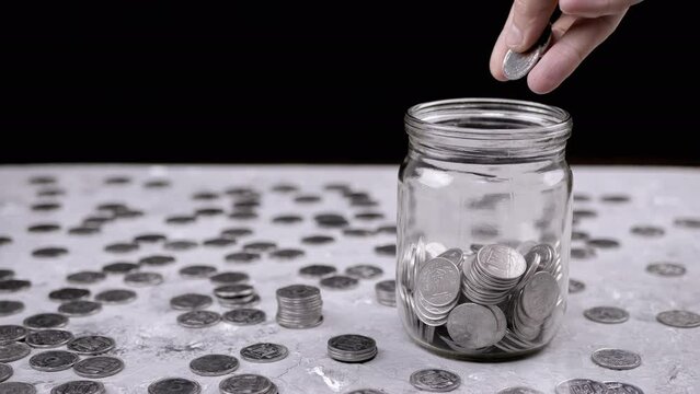 Hands Putting a Handful of Accumulated Silver Coins into a Glass Jar. Piggy bank. Pile of scattered Ukrainian five kopecks on the table. Black background. Financial crisis, budget savings. Poverty.