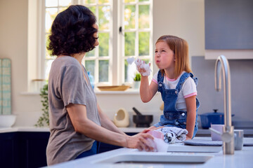 Family With Daughter Helping Older Mother To Do Washing Up In Kitchen At Home Blowing Soap Bubbles