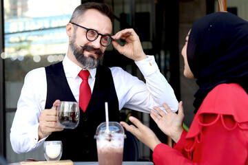 Senior elderly mature business man having a coffee at outdoor coffee shop and talking with headscarf hijab Muslim women, happy harmony people take a break from drinking beverage, enjoy relaxing.