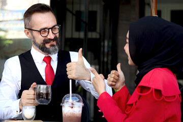 Senior elderly mature business man talking and give thumbs up with hijab Muslim women during having a coffee at outdoor cafe, happy harmony people take a break from drinking beverage, enjoy relaxing.
