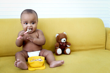 Cute little girl African American baby daughter trying to feed herself with spoon of food, infant...