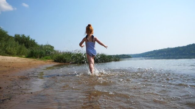 Child's bare feet run along the sandy beach by the river. The redhead girl is running on the water.