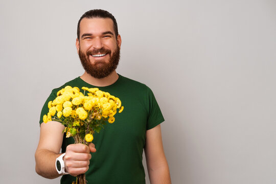 Handsome bearded man is holding a bunch of yellow flowers giving the to the camera.