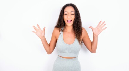 Emotive teen girl with curly hair wearing grey sport set over white background laughs loudly, hears funny joke or story, raises palms with satisfaction,