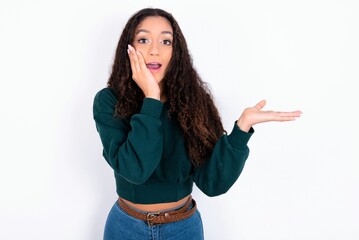 Positive glad teen girl with curly hair wearing green sweater over white background says: wow how exciting it is, indicates something.  One hand on his head and pointing with other hand.