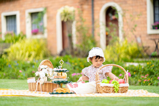 Asian Cute Little Girl Sitting On A Picnic Mat And Selecting Fruits On A Basket In Sunny Day.