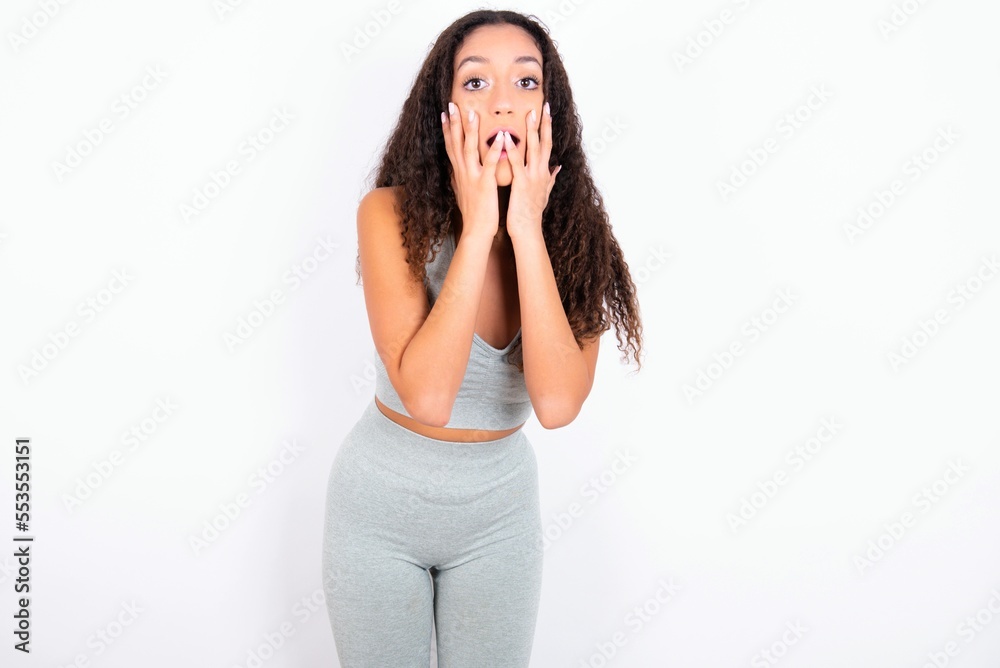 Wall mural Scared terrified Beautiful teen girl with curly hair wearing green sport set over white background shocked with prices at shop, People and human emotions concept - Wall murals