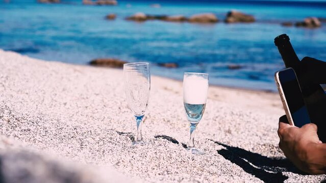 Blogger girl pours sparkling wine into glass on beach and shoots on mobile phone