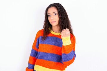 beautiful teen girl wearing striped knitted sweater over white background shows fist has annoyed face expression going to revenge or threaten someone makes serious look. I will show you who is boss