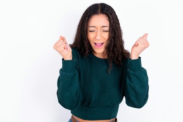 teen girl wearing knitted green sweater over white background rejoicing his success and victory clenching fists with joy being happy to achieve aim and goals. Positive emotions, feelings.