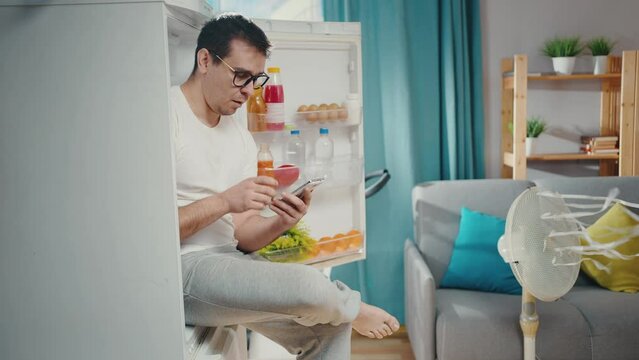 man with a smartphone in his hands searching the Internet suffering from a heat problem is sitting in the refrigerator