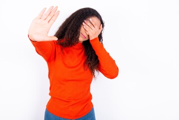 teen girl wearing knitted red sweater over white background covers eyes with palm and doing stop...