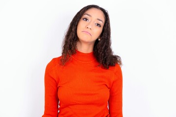Gloomy, bored teen girl wearing knitted red sweater over white background frowns face looking up, being upset with so much talking hands down, feels tired and wants to leave.