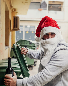 Papa Noel sin techo bebiendo vino y urgando en los contenedores de basura