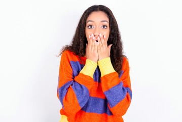 beautiful teen girl wearing knitted colourful sweater over white background keeps hands on mouth,...