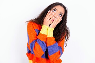 beautiful teen girl wearing knitted colourful sweater over white background  keeps hands on cheeks...