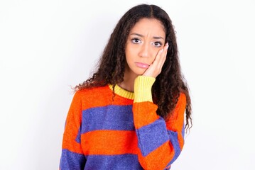 Sad lonely beautiful teen girl wearing knitted colourful sweater over white background touches cheek with hand bites lower lip and gazes with displeasure. Bad emotions